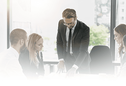 Business professional standing and presenting to colleagues seated around table in modern office with large windows