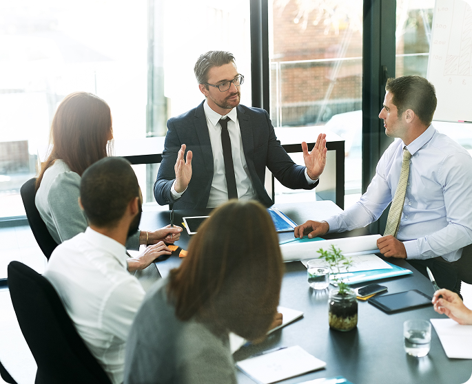Business leader in suit leading team discussion around conference table with laptops, documents, and charts in modern office.