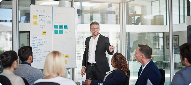 Team meeting in a modern office, gesturing toward colleagues while standing beside a whiteboard with workflow notes